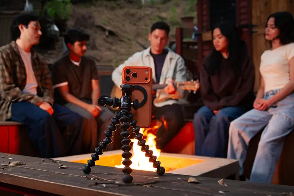 Flexible tripod supporting a smartphone on a wooden surface, facing friends gathered around a fire pit