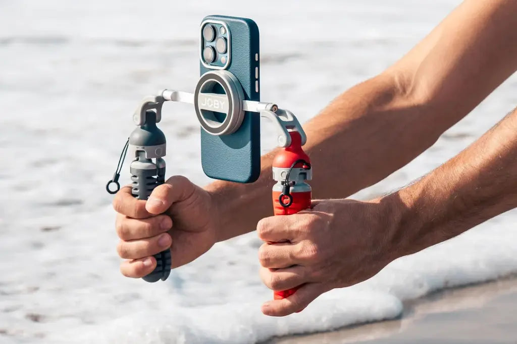 Person holding a smartphone vertically on a JOBY dual-grip rig at the beach.